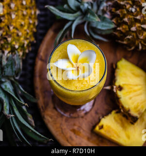 Un bicchiere di fresco succo di ananas decorata con fiori di Plumeria, due intere Ananas e albero grande pezzi tagliati su una rotonda vassoio in legno Foto Stock