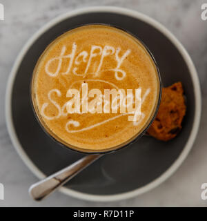 Tazza di caffè latte in nero coppa in ceramica su un tavolo mentre, con felice sabato latte art e un cookie al cioccolato in un cucchiaio sul lato. Immagine quadrata. Foto Stock