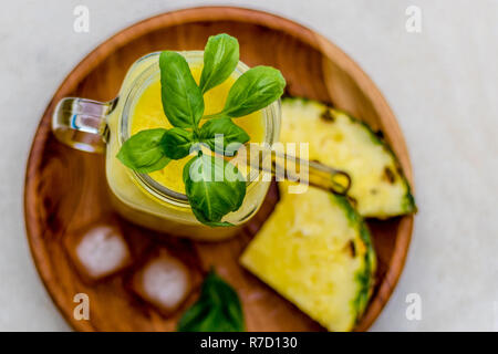 Bicchiere di fresco succo di ananas con vetro giallo paglia, sul legno scuro piastra, servita con due pezzi di ananas, cubetti di ghiaccio e le foglie di basilico. Foto Stock