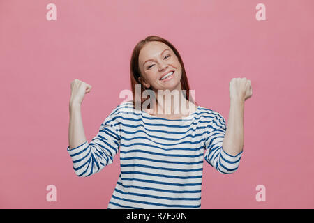 La gente, il successo, la realizzazione e la determinazione del concetto. Felice contentissimo bella donna alza le mani in pugni, celebra la sua vittoria, indossa coagulo con striping Foto Stock