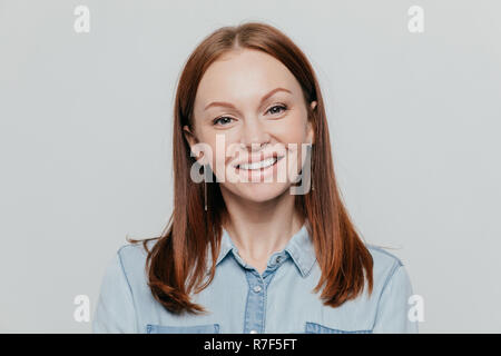 Foto di attraente giovane donna con capelli neri dritti, sorride felicemente, vestito in jeans e maglietta pone su sfondo bianco, esprime emoti positivo Foto Stock