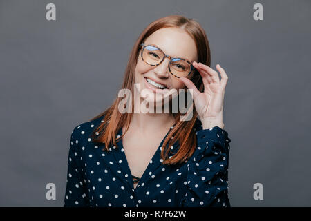 Donna sorridente con piacere l'espressione del viso, mantiene la mano sul bordo di occhiali, indossa il nero polka dot camicetta, esulta la promozione al lavoro, pone su gr Foto Stock