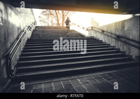 Uomo turistico in una cappa con uno zaino grande salite le scale all'alba in un ambiente urbano. Turismo invernale in concetto di città Foto Stock