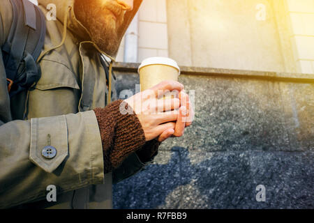 Bicchiere di carta di caffè caldo nella mano di un uomo barbuto in una camicia e un maglione marrone. Warming drink nel freddo inverno. Tenere lontano il concetto di caffè. Foto Stock