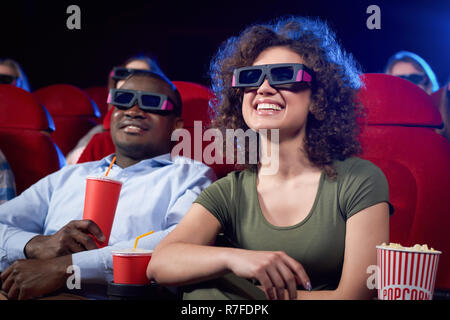 Felice carino coppia internazionale mangiare popcorn e ridere divertente commedia in cinema teatro. Ragazza attraente e bello afro man aventi data romantico e godendo interessante filmato. Foto Stock