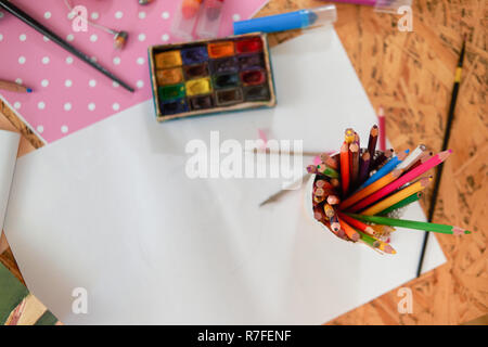 Contenitore di matite colorate sul foglio di carta bianco. La tabella con il pittore di forniture. Tavolozza di acquerelli, spazzole e foglio di pois rosa carta. Foto Stock