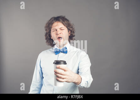 Stanco guy sbadigli con sonnolenta di espressione e la bocca spalancata, trattiene la carta monouso tazza di caffè, vestito in maglietta blu e il filtro bow tie pone contro uno sfondo grigio. Mattina tempo. Pausa caffè concept Foto Stock