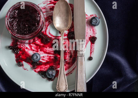 Close-up di un vasetto con confettura di mirtilli, posate. Tutto su un velluto scuro dello sfondo. Foto Stock