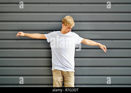 Modello urbano di vestiti. Slim hipster con due bracci sollevati vicino al grigio metallo parete sagomata in un fustellato T-shirt e pantaloncini marrone. Mockup pronto Foto Stock