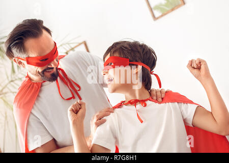 Padre e figlio in costumi superheroe a casa in piedi che guarda ad ogni altro laughign allegro Foto Stock