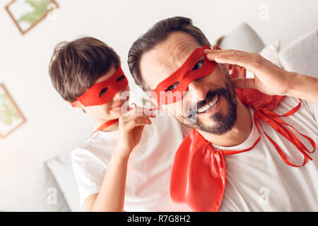 Padre e figlio in costumi superheroe a casa boy aiutando la maschera di legatura di papà sorridente close-up Foto Stock