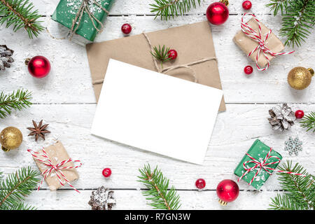 Natale bigliettino vuoto nella cornice di Abete rami, bacche rosse, decorazioni e confezioni regalo su bianco sullo sfondo di legno. mock up. flat Foto Stock