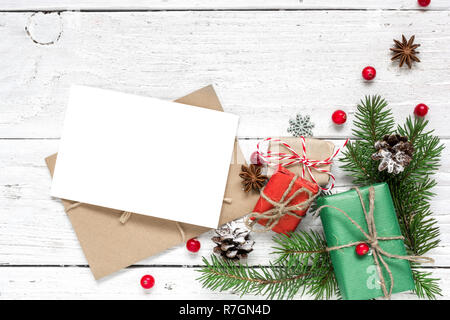 Natale bigliettino vuoto e la busta con abete rami, bacche rosse, pigne e confezioni regalo su bianco sullo sfondo di legno. mock up. flat Foto Stock