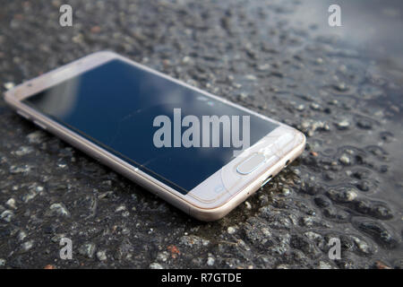 Moderne smart phone con schermo rotto vetro giacente in una pozza sul asfalto umido Foto Stock