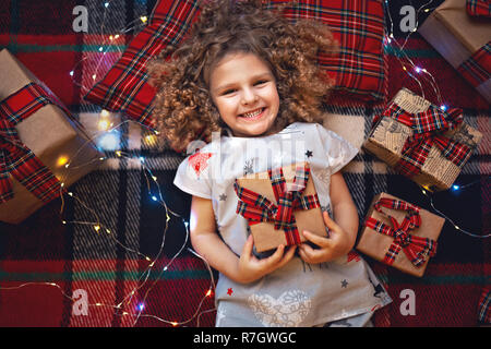 Closeup ritratto di sorridere grazioso fanciullo in vacanza natale pigiami azienda confezione regalo. Vista superiore del capretto felice posa su plaid a scacchi vicino presenta. Foto Stock