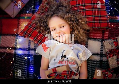 Closeup ritratto di sorridere grazioso fanciullo in vacanza natale pigiami azienda confezione regalo. Vista superiore del capretto felice posa su plaid a scacchi vicino presenta. Foto Stock
