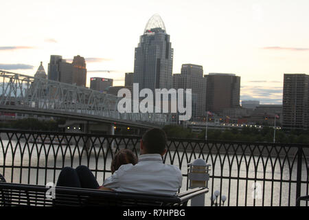 Giovane seduto su un banco di lavoro sulle rive del fiume Ohio, affacciato sul centro cittadino di Cincinnati, Ohio Foto Stock