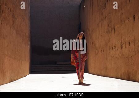 Una signora in rosso e rosa cammina verso di noi verso il basso una illuminata vividamente road, circondato da alte mura oscuro Foto Stock