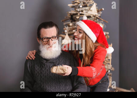 Bello barbuto senior papà indossando occhiali e maglione grigio accetta un dono dal suo giovane capelli lunghi nella figlia il maglione rosso che indossa Santa hat, egli sensazione scioccato e sorpreso, Anno nuovo, Natale, vacanze, souvenir e regali, shopping, sconti, negozi, Snow Maiden Babbo Natale,make-up, Hairstyle, carnevale. Foto Stock