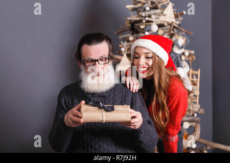 Pregi senior papà con la barba bianca che indossano occhiali e maglione grigio considerando un regalo dal suo giovane dai capelli lunghi figlia in maglione rosso indossando Santa hat, lei guarda del daddy reazione, anno nuovo, Natale, vacanze, souvenir e regali, shopping, sconti, negozi, Snow Maiden Babbo Natale,make-up, Hairstyle, carnevale. Foto Stock