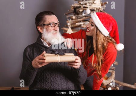 Pregi senior papà con la barba bianca che indossano occhiali e maglione grigio grazie per il dono di suoi giovani dai capelli lunghi figlia in maglione rosso indossando Santa hat, Anno nuovo, Natale, vacanze, souvenir e regali, shopping, sconti, negozi, Snow Maiden Babbo Natale,make-up, Hairstyle, carnevale. Foto Stock