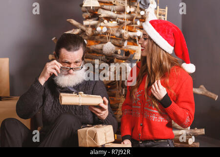 Pregi senior papà con la barba bianca che indossano occhiali e maglione grigio considerando un regalo dal suo giovane dai capelli lunghi figlia in maglione rosso indossando Santa hat, sorride e orologi del daddy reazione, anno nuovo, Natale, vacanze, souvenir e regali, shopping, sconti, negozi, Snow Maiden Babbo Natale,make-up, Hairstyle, carnevale. Foto Stock