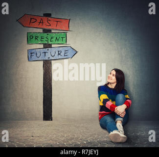 Giovane donna seduta sul pavimento guardando un cartello con le frecce che mostra il passato, il presente e il futuro. Perse nel tempo colorato cartello stradale, destino evolu Foto Stock