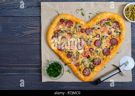 Una gustosa pizza a forma di cuore, con funghi, salame e salsiccia per pizza, olive, mais . Vista superiore Foto Stock