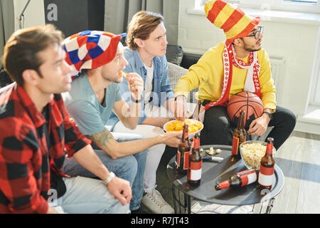 Un gruppo di giovani amici nel calcio cappelli mangiare patatine e bere birra concentrata sul match di sport in TV Foto Stock