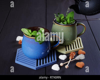 Versando il tè alla menta su un filtro con foglie di menta fresca. Due tazze, uno verde, gli altri blu. Bastoncini di zucchero e zucchero cuori come decorazione Foto Stock