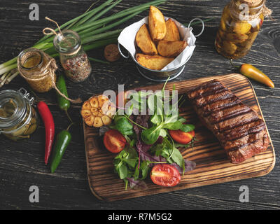 Bistecca alla griglia sulla scheda servita con erbe aromatiche, patate fritte, spezie su una superficie nera Foto Stock