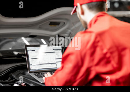 Bello meccanico automatico in rosso uniforme facendo la diagnostica motore con il calcolatore in auto di servizio Foto Stock