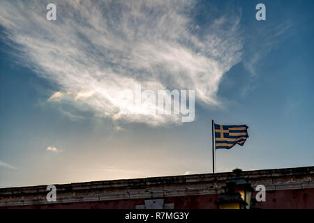 Bandiera Greca contro il sole al tramonto, scenario le nuvole e la luce proveniente da dietro la parete del fort di Corfù, Kerkira Island, Grecia. Calma spri Foto Stock