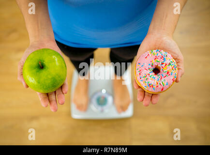 Close up della donna su scala tenendo le mani su apple e ciambella rendendo la scelta tra un sano cibo malsano dessert durante la misurazione del peso corporeo nel dado Foto Stock