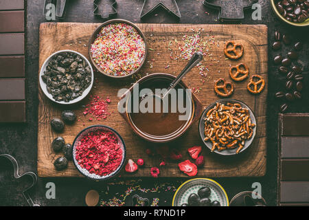 Cioccolato artigianale bar facendo. Vari condimenti e aromi ingredienti in ciotole con cioccolato fuso su dark cucina rustica sfondo tabella, Foto Stock