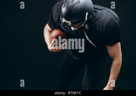 Giocatore di football americano con il casco e armatura con una sfera contro la parete nera Foto Stock