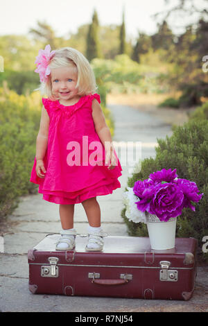 Incredibile deep blue eyes Baby girl bambino vestito elegante colorato abito rosa con Brillante peli biondi e sandali bianchi in posa per sedersi per fotocamera estate ce Foto Stock