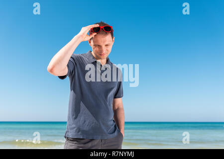 Giovane uomo hipster millenaria faccia a contatto con gli occhiali sulla spiaggia durante la giornata di sole con occhiali da sole rosso in Florida Panhandle oceano, guardando verso il basso Foto Stock