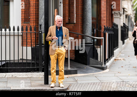 London, Regno Unito - 16 Settembre 2018: Elegante senior uomo turistica con giallo arancione pantaloni camicia di moda in piedi in attesa di cross street road fumatori ciga Foto Stock