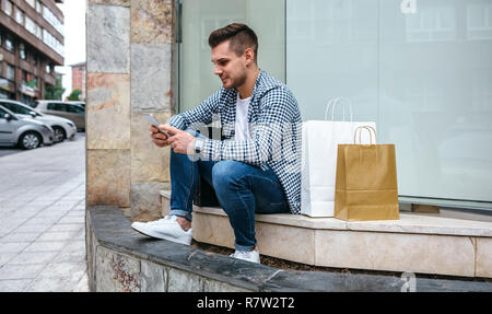 Giovane con le borse della spesa seduti sul cordolo alla vetrina di un negozio Foto Stock