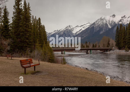 Panca seduta sulla prua riverbank vista del Fiume Bow, la Snow capped mountain e il ponte al centro Canmore nelle Montagne Rocciose Canadesi in Alberta Foto Stock