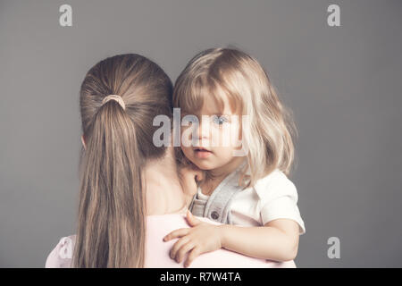 Giovani e sottile per la madre sta con la schiena e tiene fra le braccia un piccolo bambino meraviglioso. Adorabile bionda dagli occhi blu bambina guarda la fotocamera Foto Stock