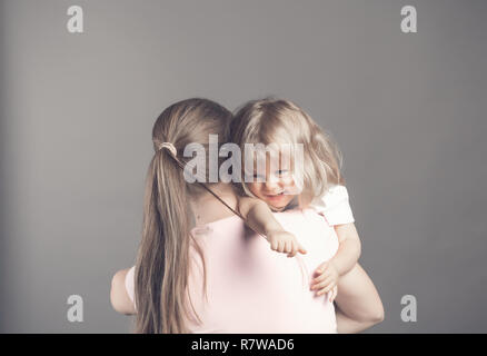 Ridere bambina sembra felice nella madre di braccia. Bellissima bambina mamma tira i capelli e sorridente. Donna che mantiene la sua figlia di stare sulla barra grigia ba Foto Stock