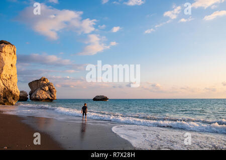 Un ragazzo scalzi gioca in spiaggia contro la Petra tou Romiou scogli immersi nella luce del pomeriggio, in Paphos, Cipro. La spiaggia è considerata essere Aphr Foto Stock