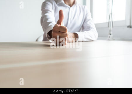 Vista frontale di imprenditore o datore di lavoro seduto alla sua scrivania in ufficio che ti mostra un pollice in alto segno. Foto Stock
