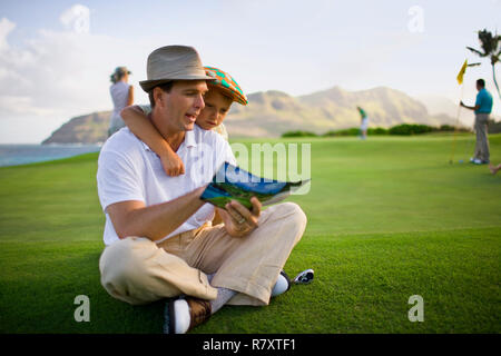 Golfista maschio si siede con le gambe incrociate sul prato con suo figlio appoggiata sopra la sua spalla per vedere la brochure del suo padre è la lettura. Foto Stock