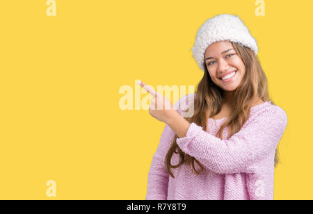 Giovane bella bruna donna che indossa un maglione e Cappello invernale su sfondo isolato allegro con un sorriso della faccia rivolta con la mano e il dito verso l'alto Foto Stock