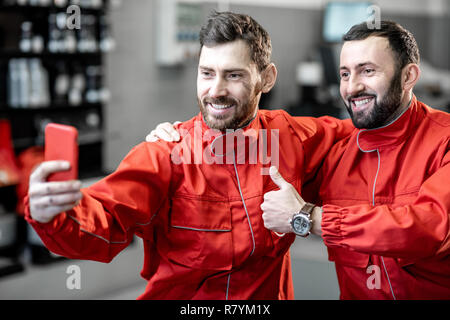 Servizio auto di lavoratori in rosso rendendo uniforme selfie foto con il telefono durante una pausa al pneumatico servizio di montaggio Foto Stock