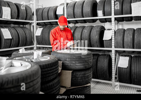 Lavoratore o venditore in rosso uniforme riempimento alcuni documenti di controllo sulle merci in magazzino con pneumatici per auto Foto Stock