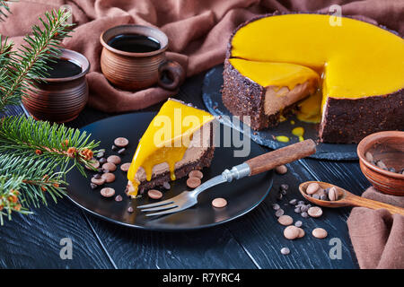 Close-up di un pezzo di cioccolato mango cheesecake servita su una piastra nera con forcella di dessert e scaglie di cioccolato. abete e tazze di caffè su un Foto Stock